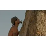 Shirtless man wearing a backwards cap looks up at a rugged rock cliff, with a small action camera mounted near his chest/torso.