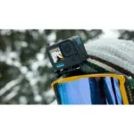 GoPro action camera mounted on a ski helmet with a snowy, blurred forest background and blue-tinted goggles nearby.