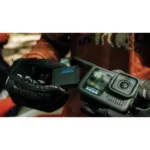 Close-up of a GoPro action camera held by gloved hands outdoors, with an orange jacket in the background.