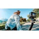 Person in a light denim jacket riding a bike while a handheld action camera is mounted on a gimbal to the right, filming the ride