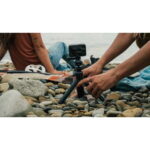 Person adjusting a small action camera on a tripod among smooth beach rocks, with another person on a blanket in the background near the water.