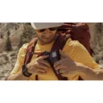 Hiker wearing a yellow shirt with a large backpack adjusts a chest-mounted action camera outdoors.