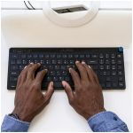 Person typing on a compact black keyboard at a white desk, wearing a blue shirt.