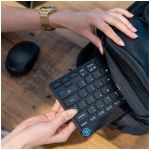 Person with a gold watch openings a backpack and removes a compact black keyboard from it on a wooden table, a wireless mouse nearby