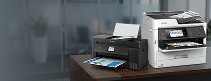 Two Epson printers on a wooden desk in an office, printing documents, with a blurred glass partition in the background.