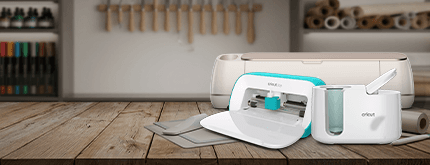 Cricut cutting machine with mats and accessories on a wooden craft table in a workshop setting.