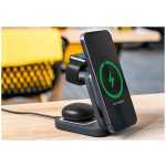 Phone on a wireless charging stand with a smartwatch charger and wireless earbuds nearby, all on a wooden table.