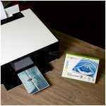 Printer on a wooden desk with a photo emerging from the slot, showing a person on a bridge in the printed photo, beside a box of photopaper kit on the right.