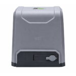 Gray compact safe with a black base and a round key lock on the front-right area. The top shows a small label, suggesting a secure storage unit.