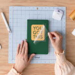 Hands peel off a clear protective sheet from a green notebook with a yellow label reading 'YOU GOT THIS' on a craft mat.