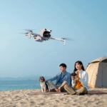 White quadcopter drone flies over a sandy beach where a couple sits on a blanket with a dog nearby and a tent in the background.