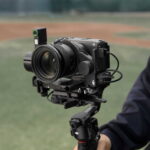 Professional camera mounted on a motorized gimbal outdoors, ready for filming; large lens facing forward with a handheld rig grip visible on the right