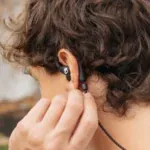 Close-up of a person wearing a black behind-the-ear headset with a small skull logo near the ear, being adjusted by a hand.
