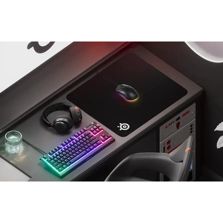 Una configuración para juegos en tonos negros con una alfombrilla SteelSeries, un ratón con un cálido brillo RGB, auriculares a juego y un teclado RGB compacto.